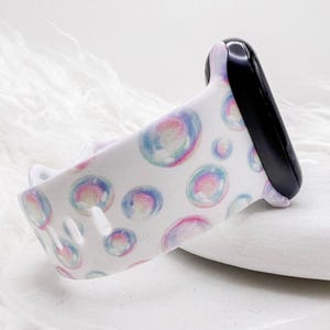 May include: A white smartwatch band featuring a colorful bubble pattern in shades of blue, pink, and purple. The band is attached to a black smartwatch face. The design is repeated across the band.