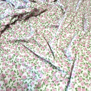 Puede incluir: Una tela blanca con un patrón denso y repetitivo de pequeñas rosas rosadas con hojas verdes. El material está fruncido y drapeado, creando pliegues y sombras suaves que resaltan la textura y el diseño floral continuo de la tela.