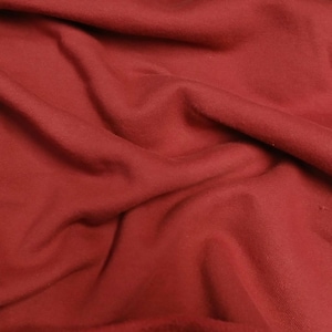May include: Close-up of a textured, red fabric with visible folds and subtle variations in tone. The material appears soft and drapes naturally, suggesting it could be used for clothing or home decor.
