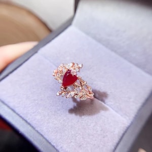 Puede incluir: Un anillo de oro rosa con una piedra de rubí en forma de pera rodeada de pequeñas piedras blancas. El anillo presenta un delicado diseño floral.