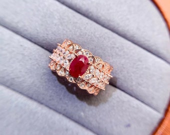May include: A rose gold ring featuring a large oval ruby surrounded by smaller white diamonds. The ring has a delicate, intricate design.