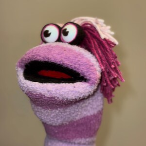 Peut inclure: Une marionnette faite à la main avec un corps rayé violet et blanc. La marionnette a de grands yeux globuleux, une grande bouche ouverte et des cheveux en fil rose. La bouche est noire avec un intérieur rouge. La marionnette est faite d'un matériau doux et duveteux.
