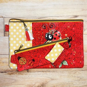 May include: Three red and yellow zippered pouches with ladybug designs. The largest pouch has a yellow polka dot fabric and the smaller pouches have a red fabric with a white script pattern and gold glitter. The pouches are on a wooden surface.