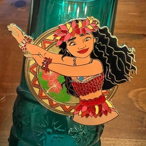 May include: A gold-toned enamel pin featuring a cartoon depiction of Moana from the Disney movie Moana. Moana is wearing a red and gold flower lei and a red and gold skirt. She has long black hair and brown eyes. The pin is set against a background of green and gold.