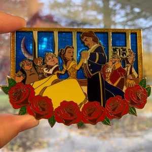 May include: A gold-toned enamel pin featuring Belle and Beast from Beauty and the Beast. The pin depicts Belle in a yellow gown and Beast in a blue suit, surrounded by roses and a stained glass window.