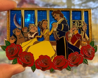 Op de afbeelding: Een gouden emaille pin met Belle en het Beest uit Belle en het Beest. De pin toont Belle in een gele jurk en het Beest in een blauw pak, omringd door rozen en een glas-in-loodraam.
