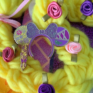 May include: Purple and glitter Minnie Mouse ears with a floral design and a purple chameleon on one ear. The ears are on a yellow background.