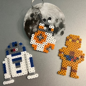 May include: Three Star Wars character keychains made of colorful perler beads. The characters are R2-D2, BB-8, and a brown figure with a yellow face.