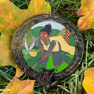 May include: A black metal pin with a green, yellow, and brown enamel design. The design features a woman with brown skin and a yellow cape playing Quidditch with a golden snitch. The pin has a black and silver crest with the letter "M" in the center.