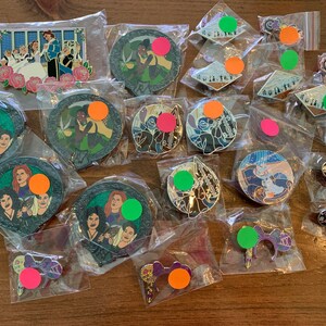 May include: A collection of enamel pins in clear plastic bags. The pins feature various designs, including characters from animated films, and decorative elements. The pins are round, shaped like keys, and other shapes. The pins have a variety of colors, including green, pink, orange, and blue.