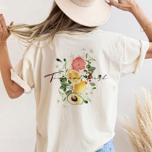 May include: A white t-shirt with a graphic of a grapefruit, lemons, and an avocado with green leaves and white flowers. The text "True & Fresh" is written in cursive on the shirt.
