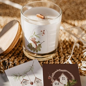 May include: Two small white houses with brown roofs are depicted on a brown card. The houses are surrounded by a brown twig wreath with a brown acorn on the left side. A green oak leaf is on the right side of the wreath. The card is on a white surface.