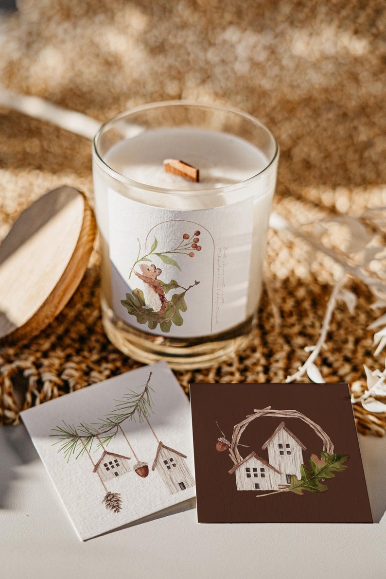 May include: Two small white houses with brown roofs are depicted on a brown card. The houses are surrounded by a brown twig wreath with a brown acorn on the left side. A green oak leaf is on the right side of the wreath. The card is on a white surface.