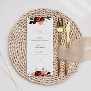May include: A white menu card with a floral design in shades of red, orange, and green. The card is placed on a woven placemat with a gold fork and knife. The text on the card reads "MENU".