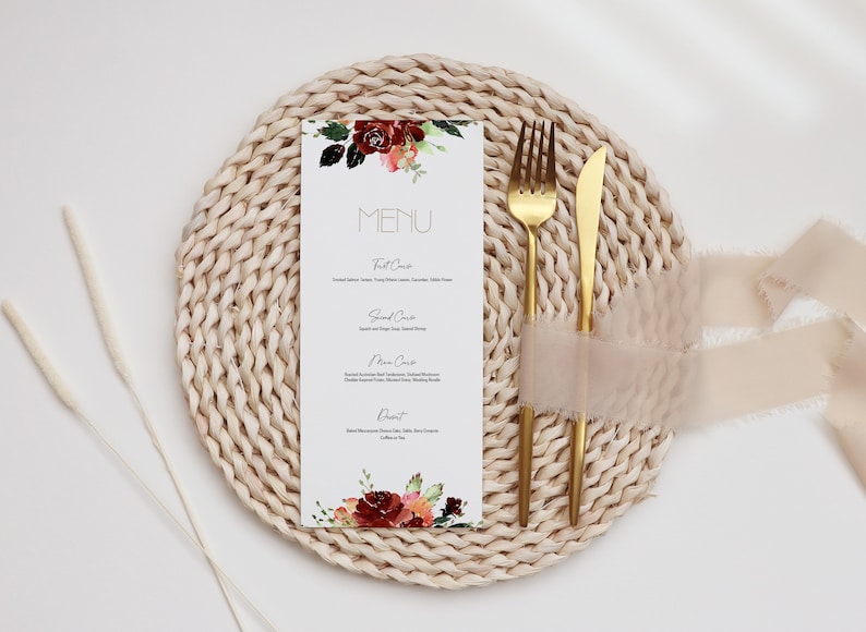 May include: A white menu card with a floral design in shades of red, orange, and green. The card is placed on a woven placemat with a gold fork and knife. The text on the card reads "MENU".