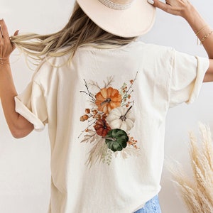 Peut inclure: Un t-shirt blanc avec un motif aquarelle de citrouilles, de feuilles et de baies. Le motif est sur le dos du t-shirt.