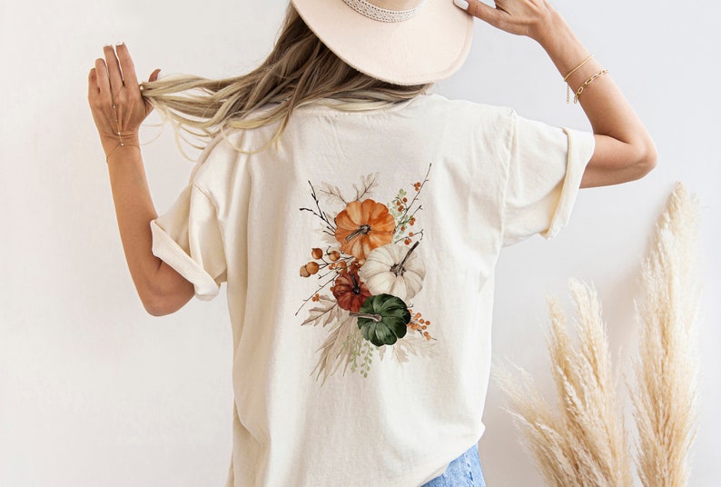 Peut inclure: Un t-shirt blanc avec un motif aquarelle de citrouilles, de feuilles et de baies. Le motif est sur le dos du t-shirt.