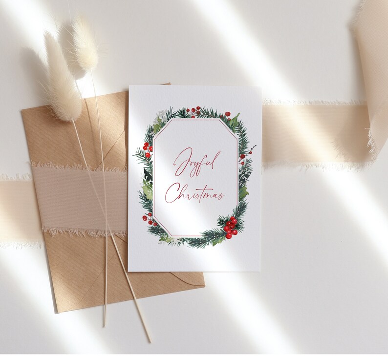 K&ouml;nnte beinhalten: Eine wei&szlig;e Karte mit einem gr&uuml;nen und roten Weihnachtskranz-Design. Der Text "Joyful Christmas" ist in roter Kursivschrift innerhalb des Kranzes geschrieben.