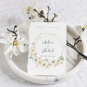 May include: Wedding invitation with a geometric gold frame and a floral wreath of white daisies. The invitation reads "Andrea & Michael" and "May 21, 2023 Cafe Flower".
