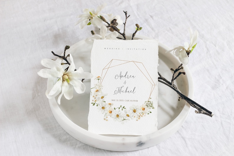 May include: Wedding invitation with a geometric gold frame and a floral wreath of white daisies. The invitation reads "Andrea & Michael" and "May 21, 2023 Cafe Flower".