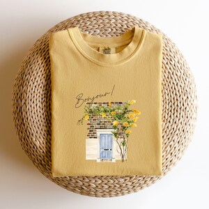 May include: A mustard yellow t-shirt with a graphic of a yellow brick building with a blue door and a vine of yellow flowers. The text "Bonjour!" is written in black above the building.