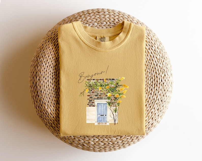 May include: A mustard yellow t-shirt with a graphic of a yellow brick building with a blue door and a vine of yellow flowers. The text "Bonjour!" is written in black above the building.