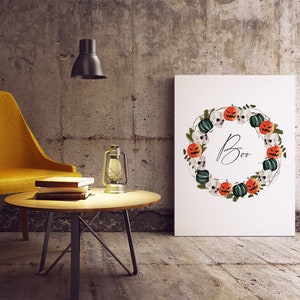 May include: A white printable art print with a Halloween wreath featuring orange and green pumpkins, skulls, and vines. The wreath surrounds the word "Boo" in black script.