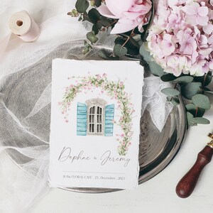 May include: A watercolor illustration of a blue window with white shutters, framed by a pink floral garland. The text "Daphne & Jeremy" and "Mille Fleurs Cafe, 21 December 2021" is printed below the illustration.