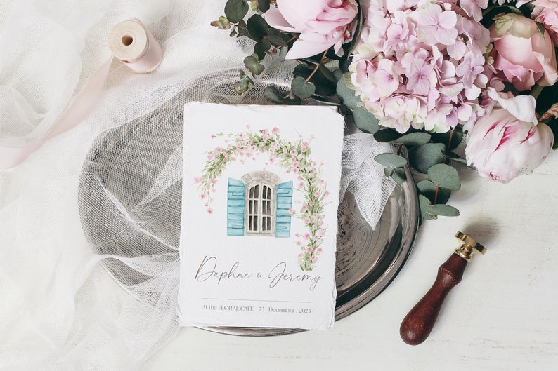 May include: A watercolor illustration of a blue window with white shutters, framed by a pink floral garland. The text "Daphne & Jeremy" and "Mille Fleurs Cafe, 21 December 2021" is printed below the illustration.