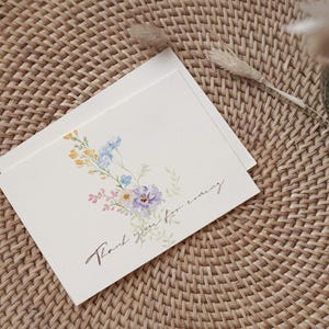 May include: Two cream-colored cards with floral watercolor illustrations and the text "Thank you for coming." The cards feature a bouquet of blue, yellow, and purple flowers. The cards are on a woven wicker surface.