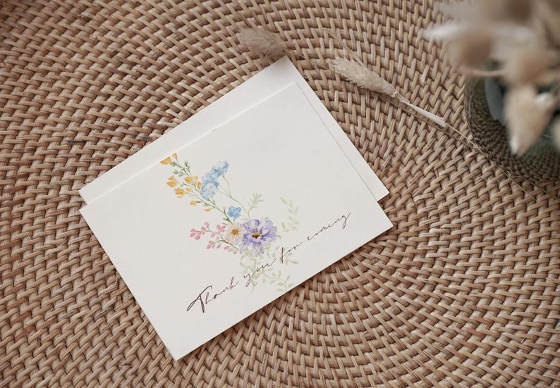 May include: Two cream-colored cards with floral watercolor illustrations and the text "Thank you for coming." The cards feature a bouquet of blue, yellow, and purple flowers. The cards are on a woven wicker surface.
