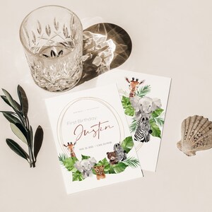 May include: Two white cards with a watercolour illustration of jungle animals, including a giraffe, elephant, and zebra. The cards are for a first birthday, with the name "Justin" and date printed. A clear glass and a seashell are also present.
