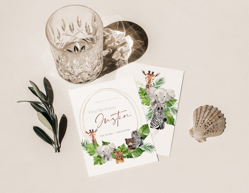 May include: Two white cards with a watercolour illustration of jungle animals, including a giraffe, elephant, and zebra. The cards are for a first birthday, with the name "Justin" and date printed. A clear glass and a seashell are also present.