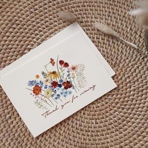 May include: Two white cards with a watercolor floral design featuring red, yellow, blue, and orange flowers. The text "Thank you for coming" is written in a cursive font. The cards are on a woven, light brown surface.