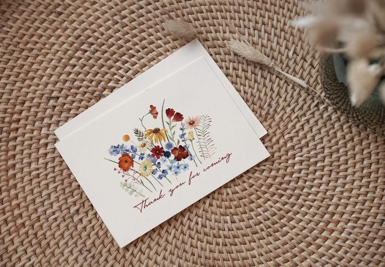 May include: Two white cards with a watercolor floral design featuring red, yellow, blue, and orange flowers. The text "Thank you for coming" is written in a cursive font. The cards are on a woven, light brown surface.
