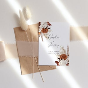 Peut inclure: Une invitation de mariage blanche avec un motif floral compos&eacute; de pampas s&eacute;ch&eacute;es, d'hortensias blancs et de roses orange. L'invitation indique "Daphne & Jeremy Au Floral Cafe 23. D&eacute;cembre 2023."