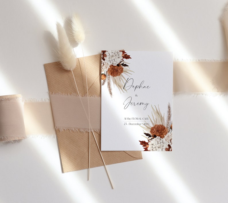 Peut inclure: Une invitation de mariage blanche avec un motif floral compos&eacute; de pampas s&eacute;ch&eacute;es, d'hortensias blancs et de roses orange. L'invitation indique "Daphne & Jeremy Au Floral Cafe 23. D&eacute;cembre 2023."