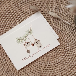 May include: A white card with a watercolor illustration of two small houses hanging from a pine branch. The text "Thank you for coming" is written in red cursive on the card.