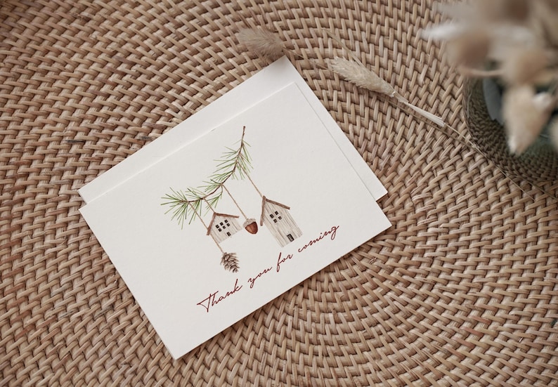 May include: A white card with a watercolor illustration of two small houses hanging from a pine branch. The text "Thank you for coming" is written in red cursive on the card.