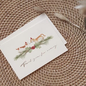 May include: Christmas greeting cards featuring a watercolor illustration of a dog and a cat wearing Santa hats, with a sprig of holly and the text "Thank you for coming." The cards are on a woven surface.