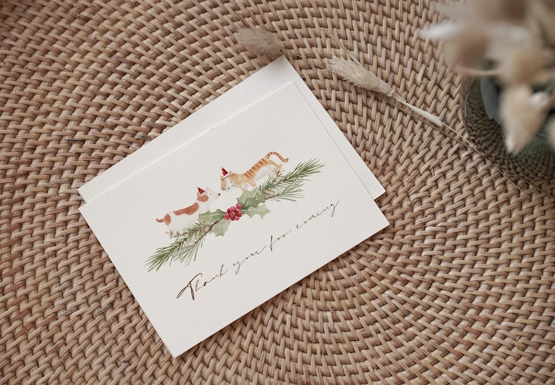 May include: Christmas greeting cards featuring a watercolor illustration of a dog and a cat wearing Santa hats, with a sprig of holly and the text "Thank you for coming." The cards are on a woven surface.