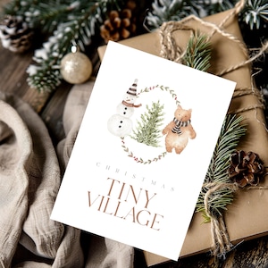 May include: A Christmas card with a watercolor illustration of a snowman, a bear, and a Christmas tree inside a wreath. The card reads "CHRISTMAS TINY VILLAGE" in brown text. The card is on a wrapped gift.