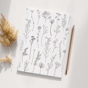 May include: A white notebook featuring a black and white line drawing of assorted flowers and plants. The notebook is lying flat on a white surface.