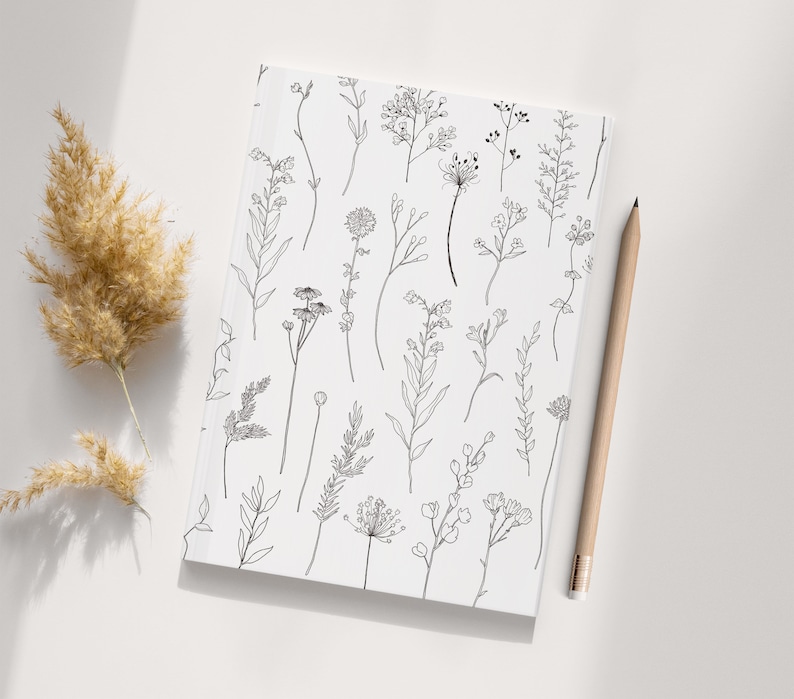 May include: A white notebook featuring a black and white line drawing of assorted flowers and plants. The notebook is lying flat on a white surface.