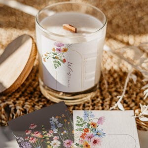 May include: A white candle with a wooden wick in a glass jar. The candle has a floral label with the text "Wildflower" and a watercolor illustration of wildflowers. The candle is sitting on a woven surface with two small cards with floral illustrations and the text "Wildflower" on them.