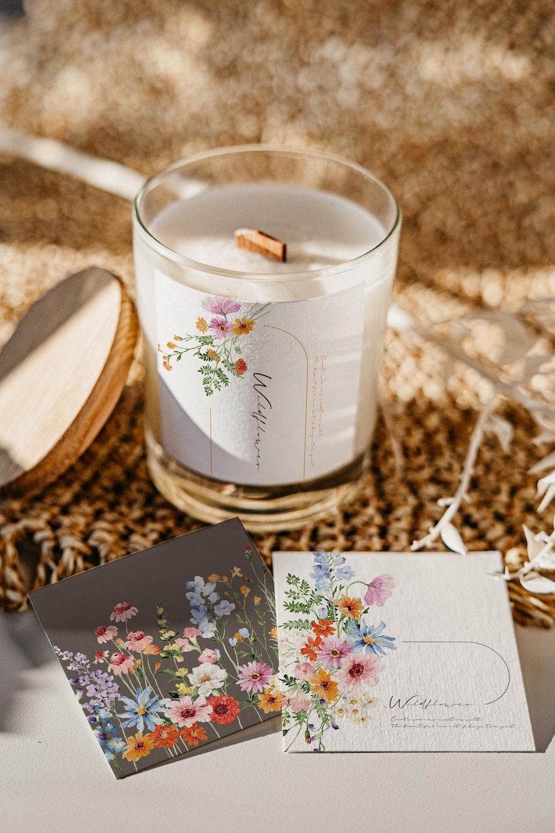 May include: A white candle with a wooden wick in a glass jar. The candle has a floral label with the text "Wildflower" and a watercolor illustration of wildflowers. The candle is sitting on a woven surface with two small cards with floral illustrations and the text "Wildflower" on them.