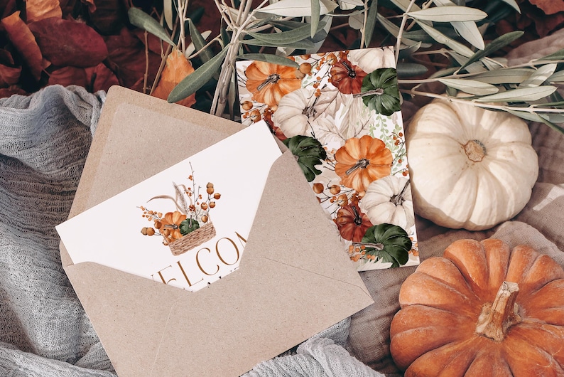 May include: A brown envelope with a white card inside. The card has a watercolor illustration of a basket filled with pumpkins and fall foliage. The text "Welcome" is printed on the card. The envelope is sitting on a brown fabric surface with a white pumpkin and an orange pumpkin in the background.
