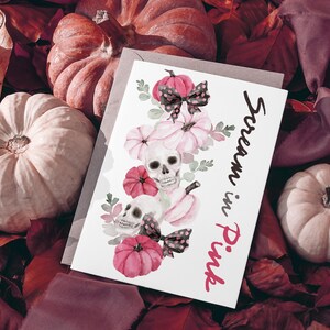 May include: A greeting card with the text "Scream in Pink" features watercolor illustrations of pink pumpkins, skulls, and bows. The card is set against a backdrop of pumpkins and fall foliage, creating a spooky yet feminine aesthetic.