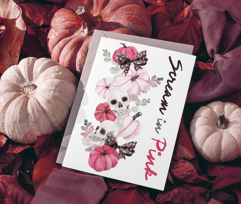 May include: A greeting card with the text "Scream in Pink" features watercolor illustrations of pink pumpkins, skulls, and bows. The card is set against a backdrop of pumpkins and fall foliage, creating a spooky yet feminine aesthetic.