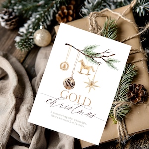 May include: A white greeting card featuring a watercolor illustration of a pine branch with hanging gold ornaments including a rocking horse, a star, and a round bauble. The text reads GOLD christmas, A season wrapped in golden light, filled with warmth and wonder.